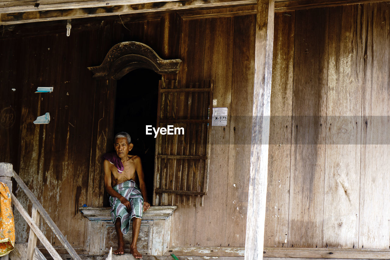 Full length of shirtless man sitting by wooden house