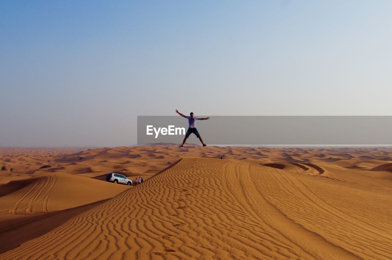 Man with arms outstretched jumping on desert against sky during sunset