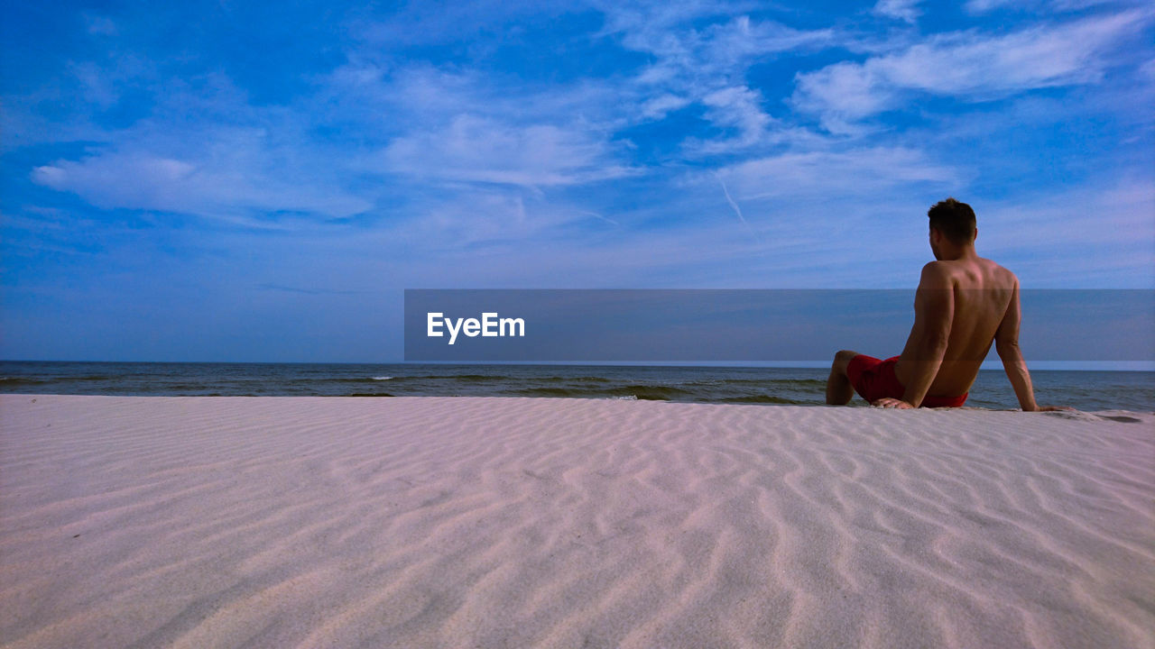 Rear view of shirtless man sitting at sandy beach against cloudy sky