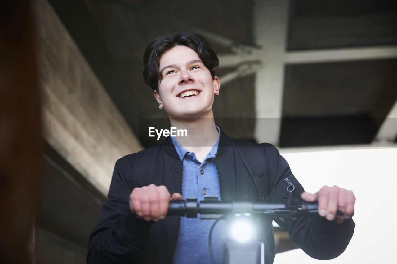 Smiling teenage boy with electric push scooter looking away while standing below bridge