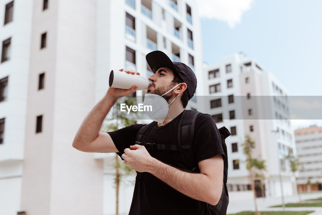 Low angle of male in respiratory mask and black wear drinking water from bottle near buildings and looking away