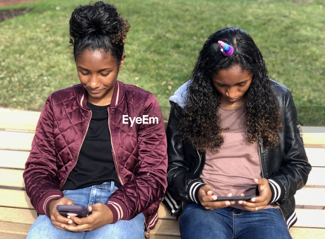 Female friends using mobile phones while sitting on bench in park