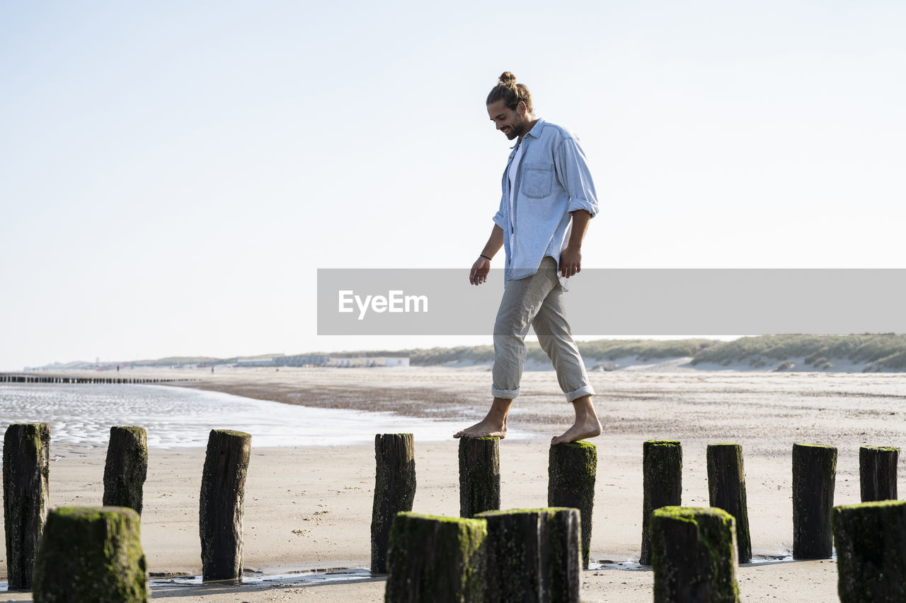 Smiling young man walking on wooden posts at beach against clear sky