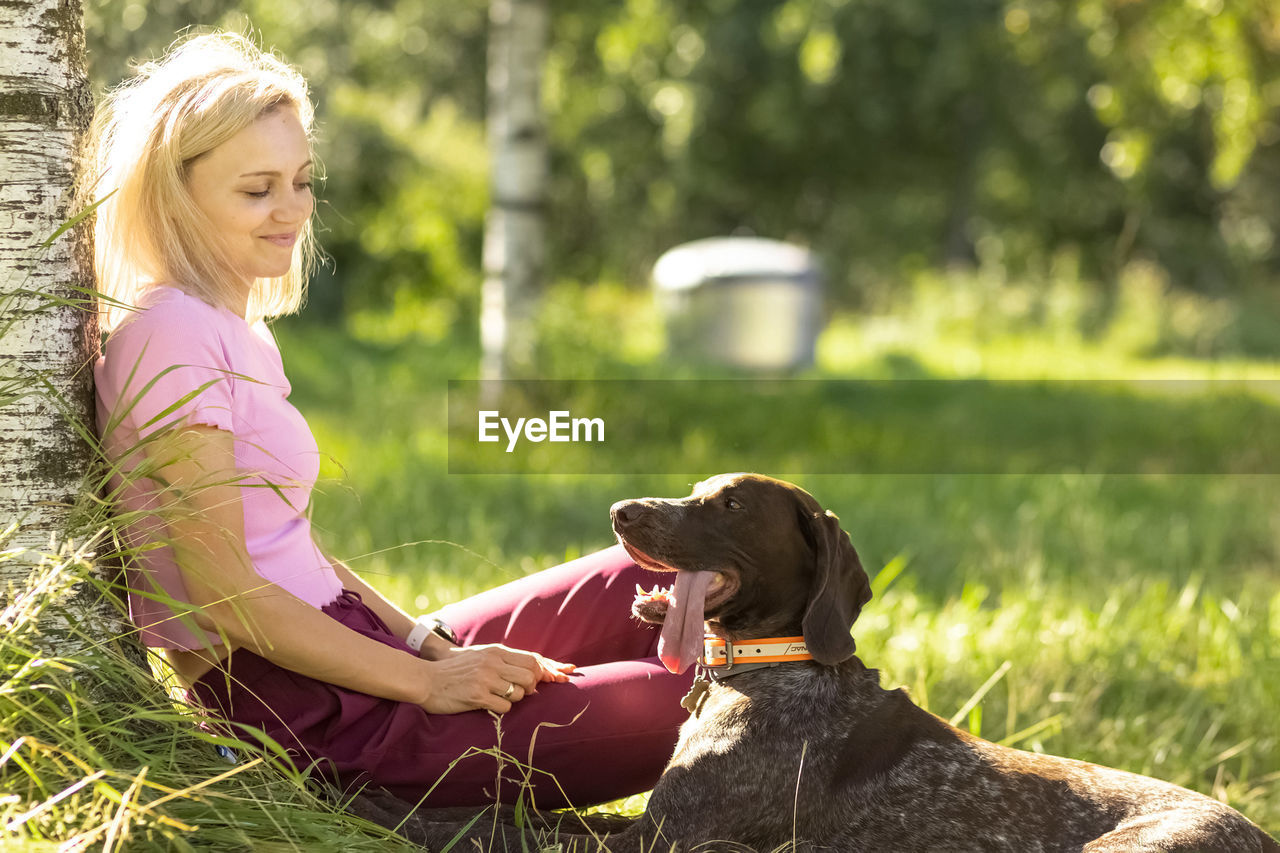 Portrait of a young blonde woman sitting,relaxing  near a tree in the park with a hunting dog