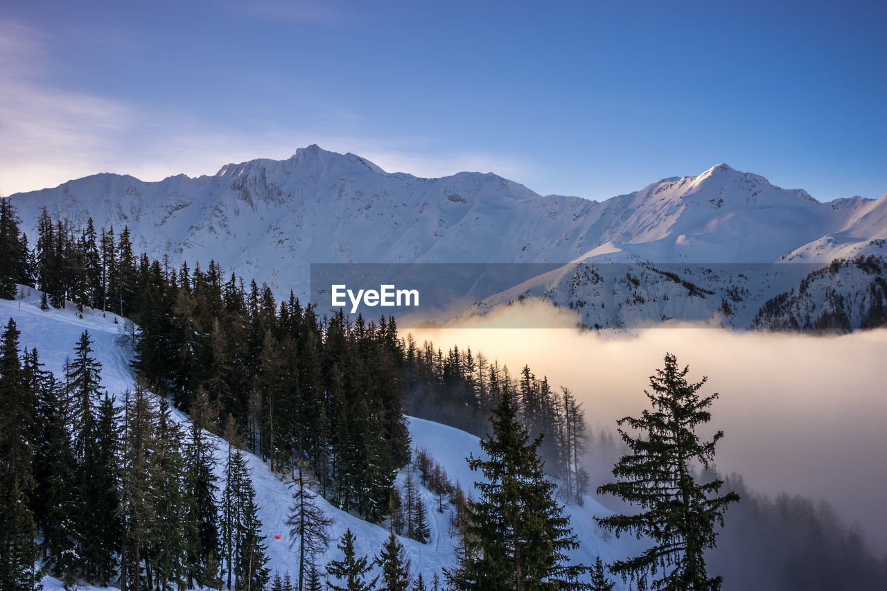 The sun shine down on a cloud filled valley, surrounded by snow covered mountains