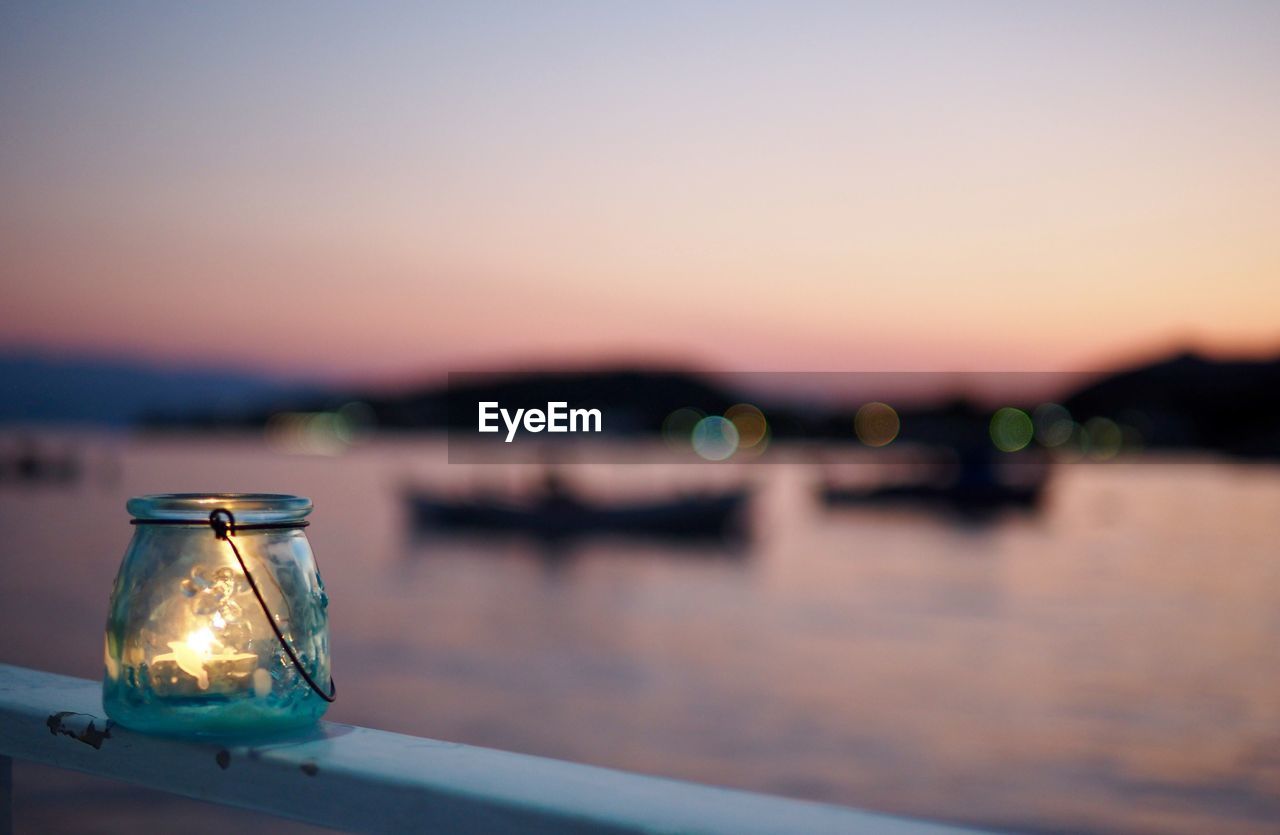 Lit candle in glass jar on railing by river against clear sky during sunset