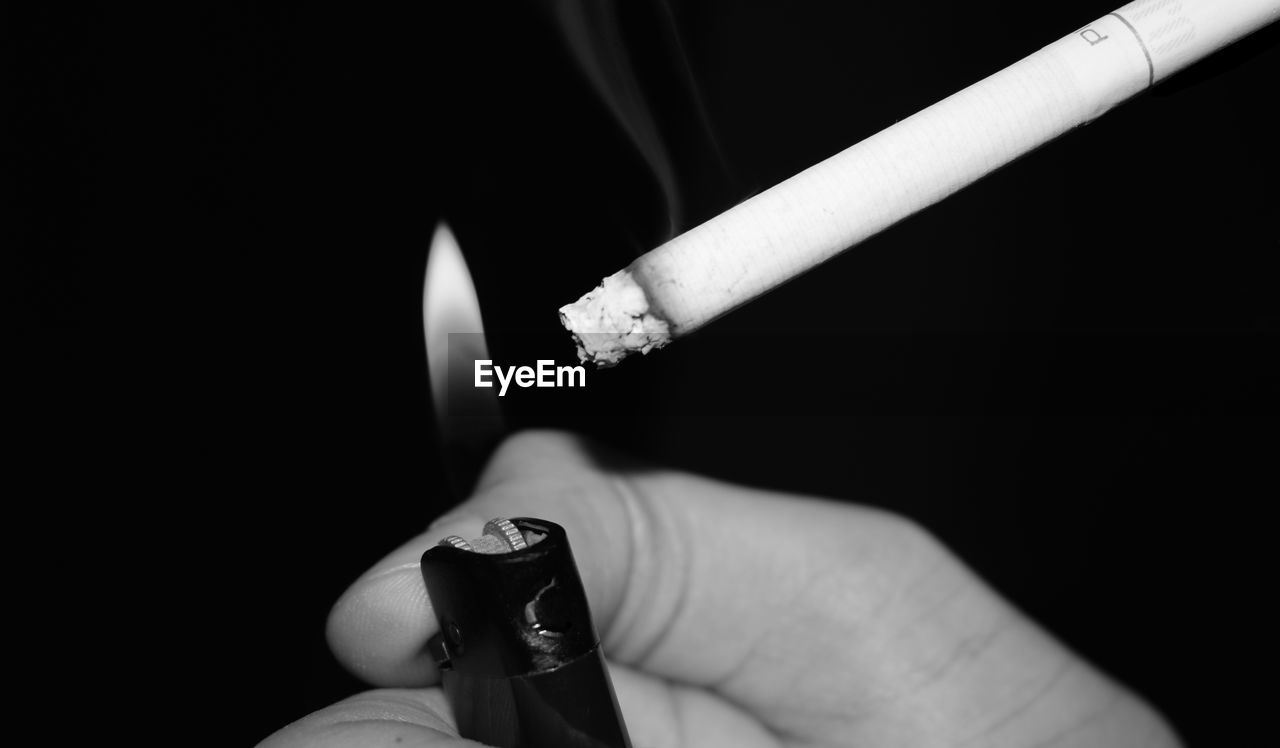 Close-up of hand holding burning lighter by cigarette against black background
