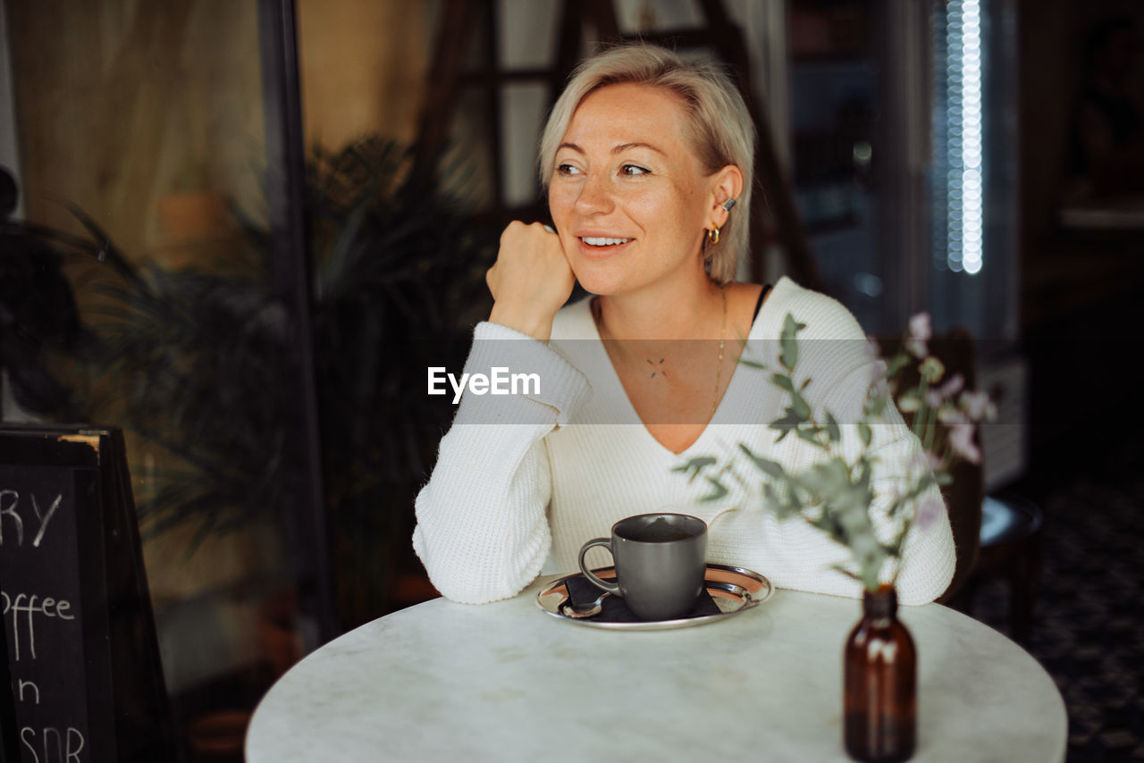 Blond woman smiling while sitting at table in cafe with cup of coffee