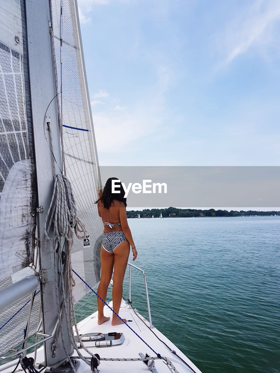 Rear view of woman wearing bikini standing on sailboat in sea against sky