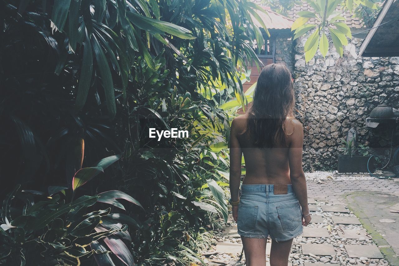Rear view of young shirtless woman walking by plants