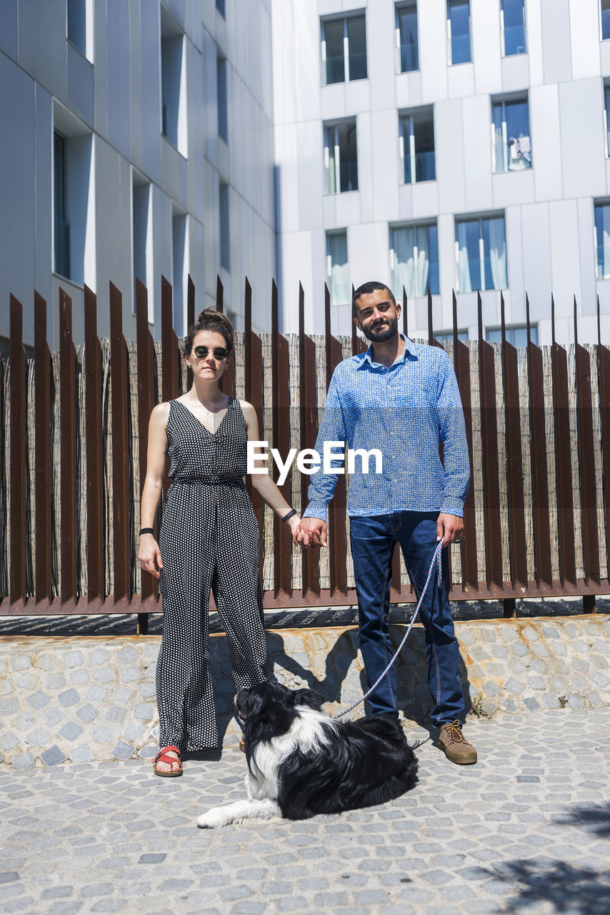 Full length portrait of couple with dog holding hands against building