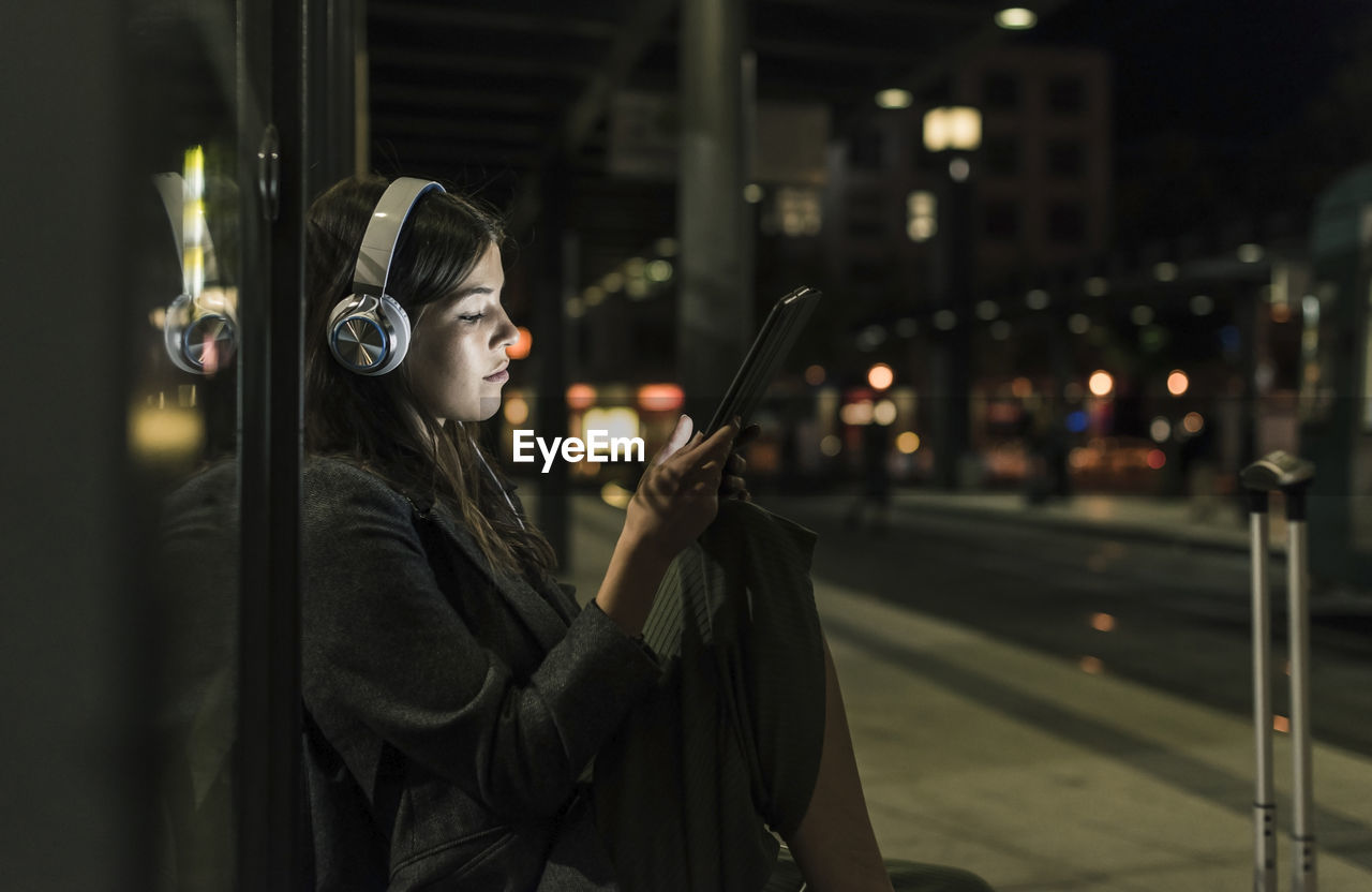 Young woman with headphones waiting at the station by night using tablet