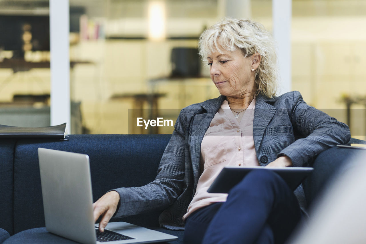 Businesswoman using laptop and digital tablet on sofa at office lobby