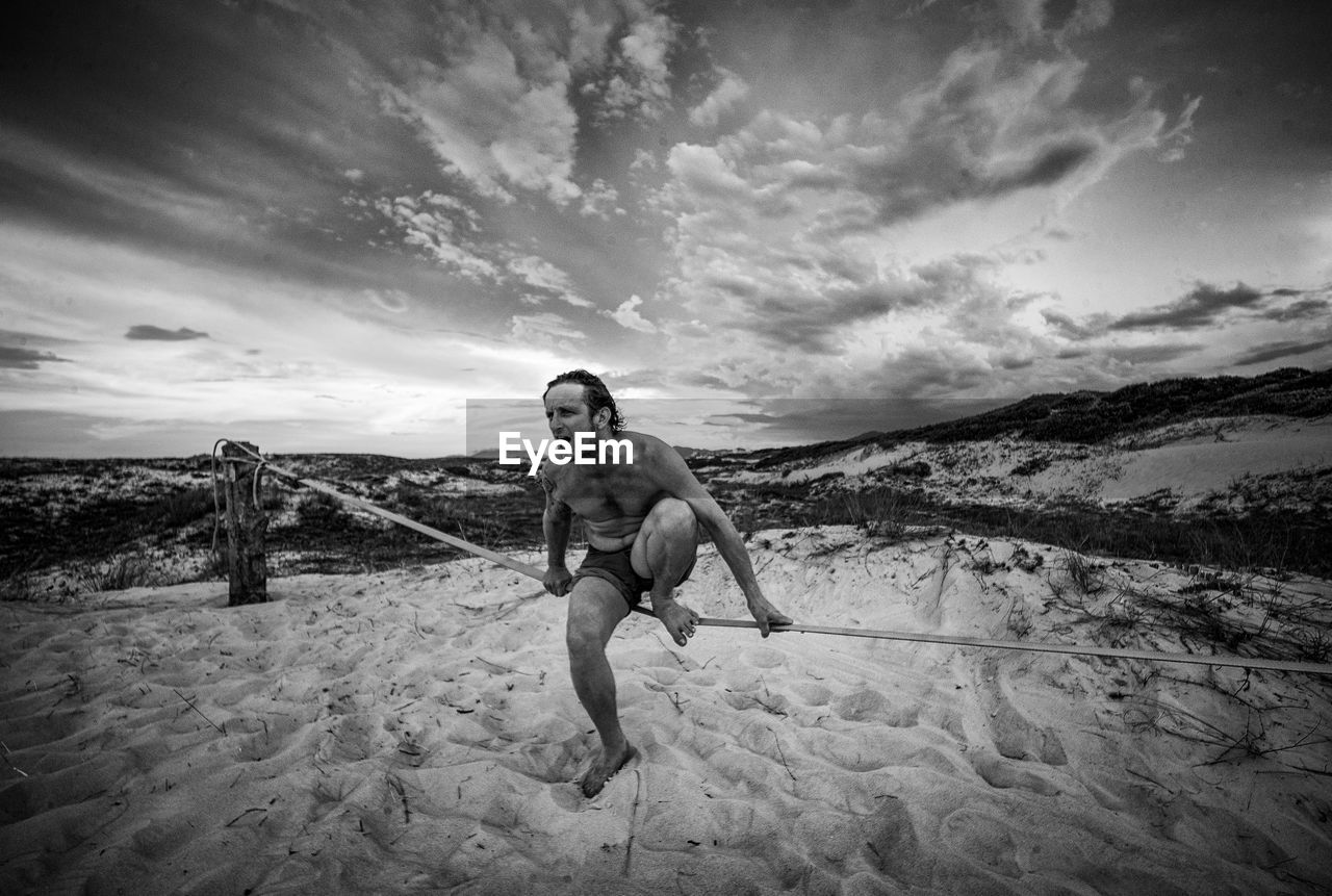 Shirtless man sitting on rope fence at beach against sky