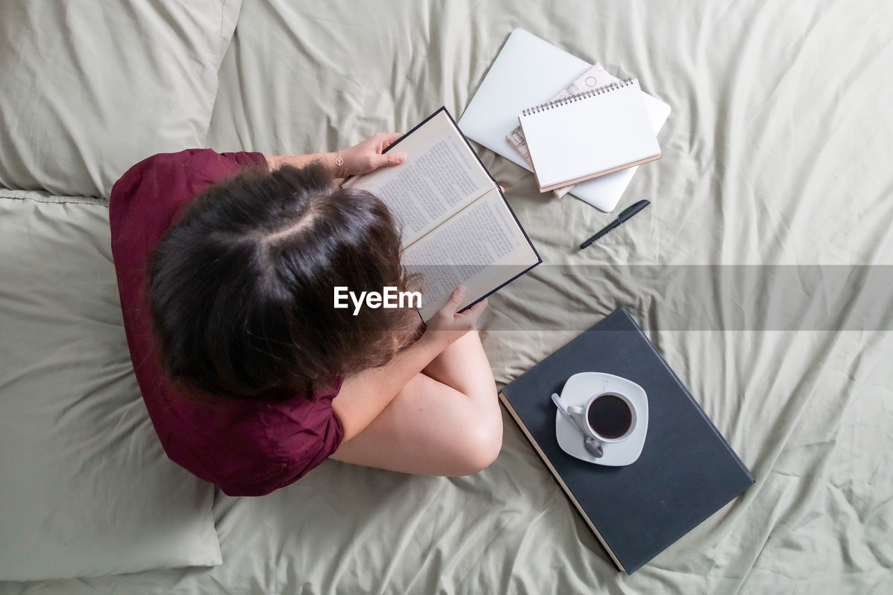 High angle view of woman reading book on bed while having coffee at home