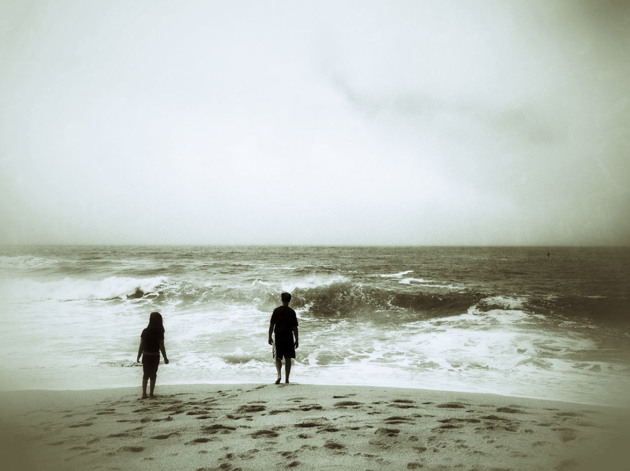 Full length rear view of people standing in front of wave on beach against sky