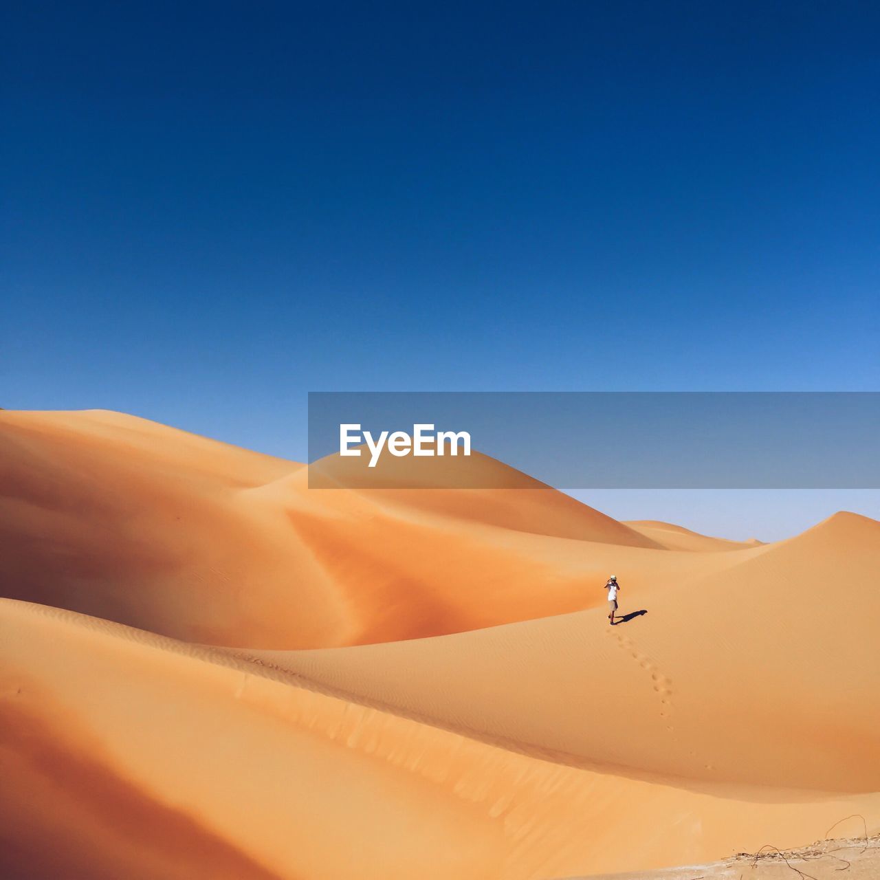 Distant view of man walking on sand dune in desert against clear blue sky