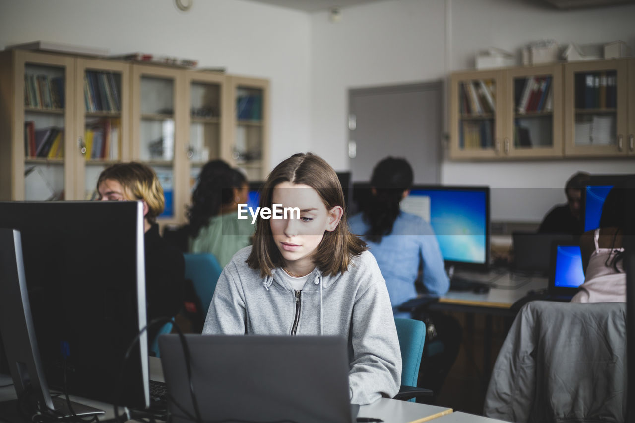 Confident female student using laptop at desk against teacher and friends sitting in computer lab