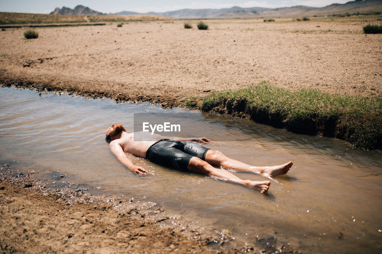 SIDE VIEW OF SHIRTLESS MAN LYING IN WATER AT SHORE