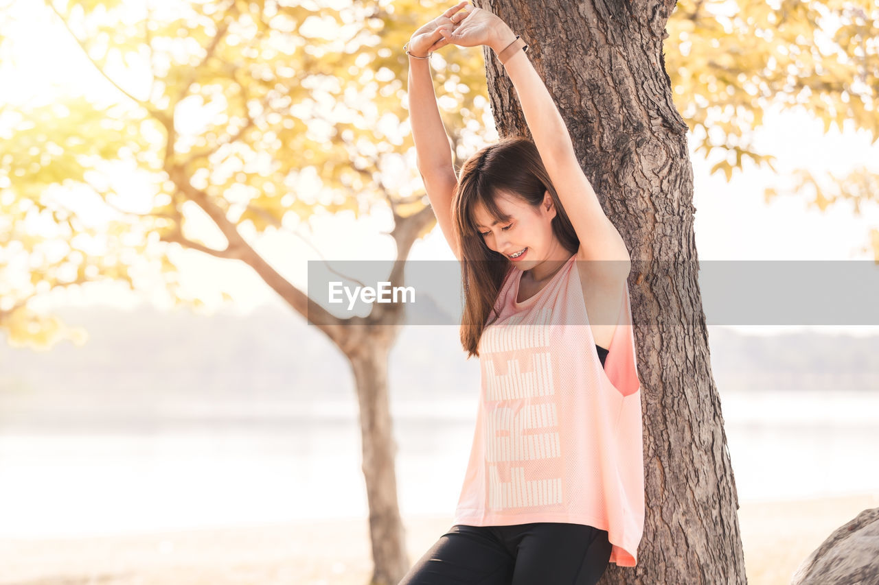 Young woman with arms raised standing against tree trunk