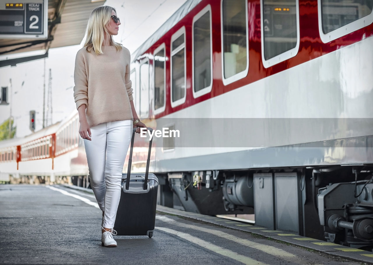 Blonde woman with her luggage go and speak in her mobile near red train rail station under sun light