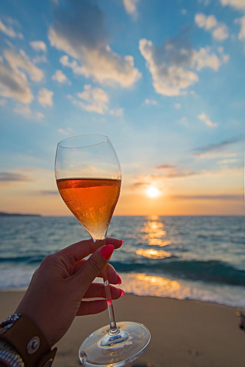 Cropped hand holding champagne flute at beach against sky during sunset