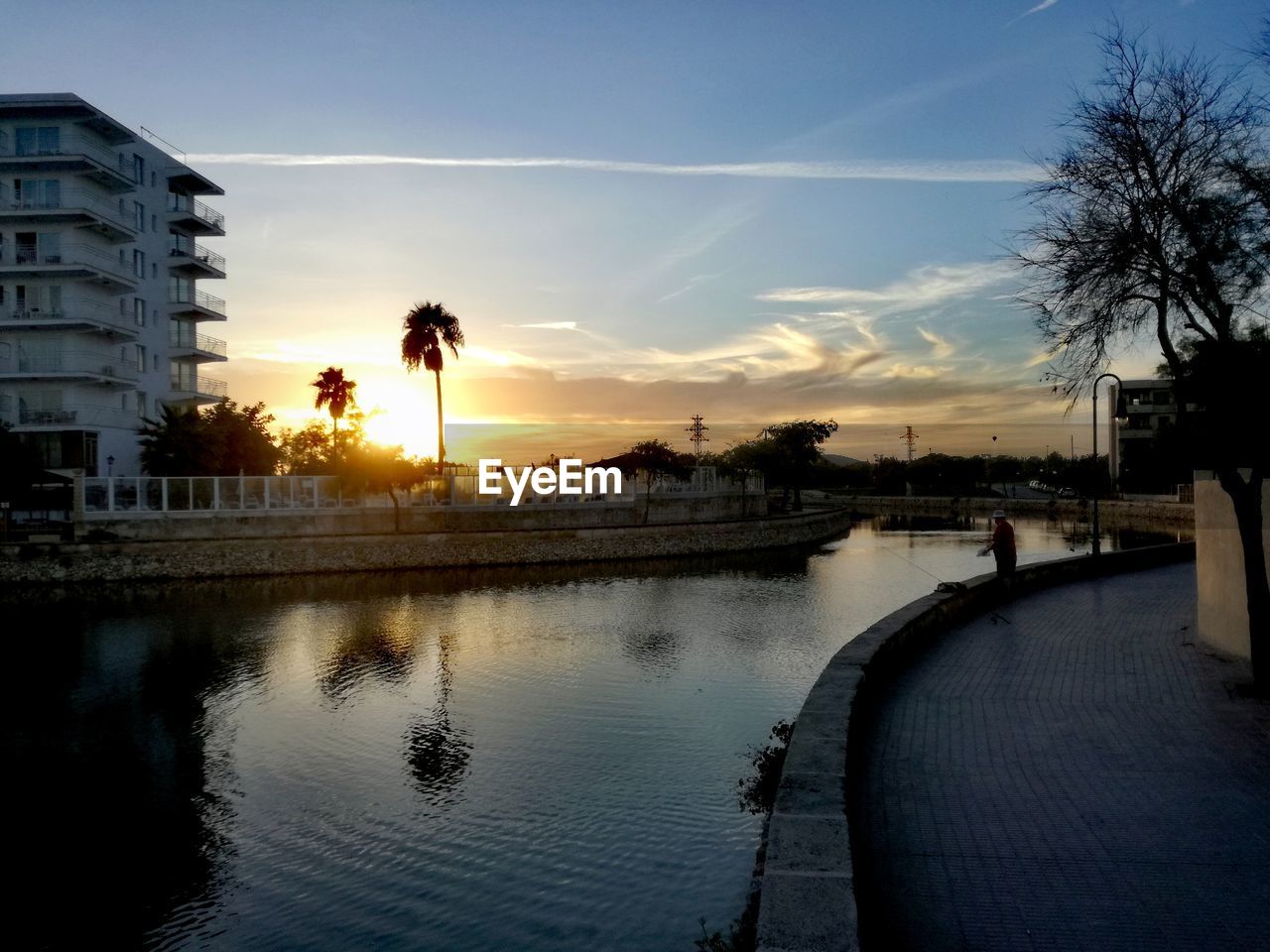 SILHOUETTE BUILDINGS BY RIVER AGAINST SKY DURING SUNSET