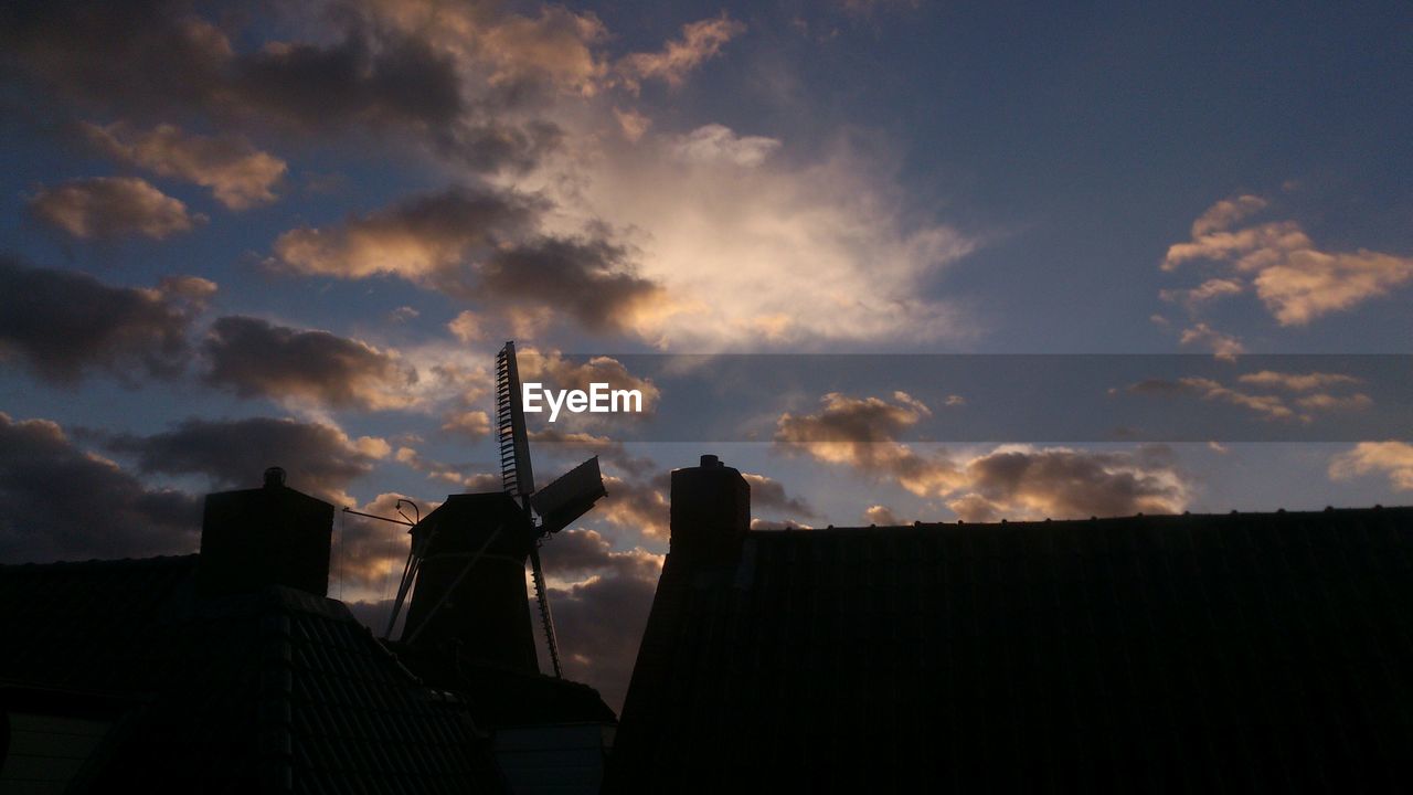 Silhouette traditional windmill by houses against sky during sunset