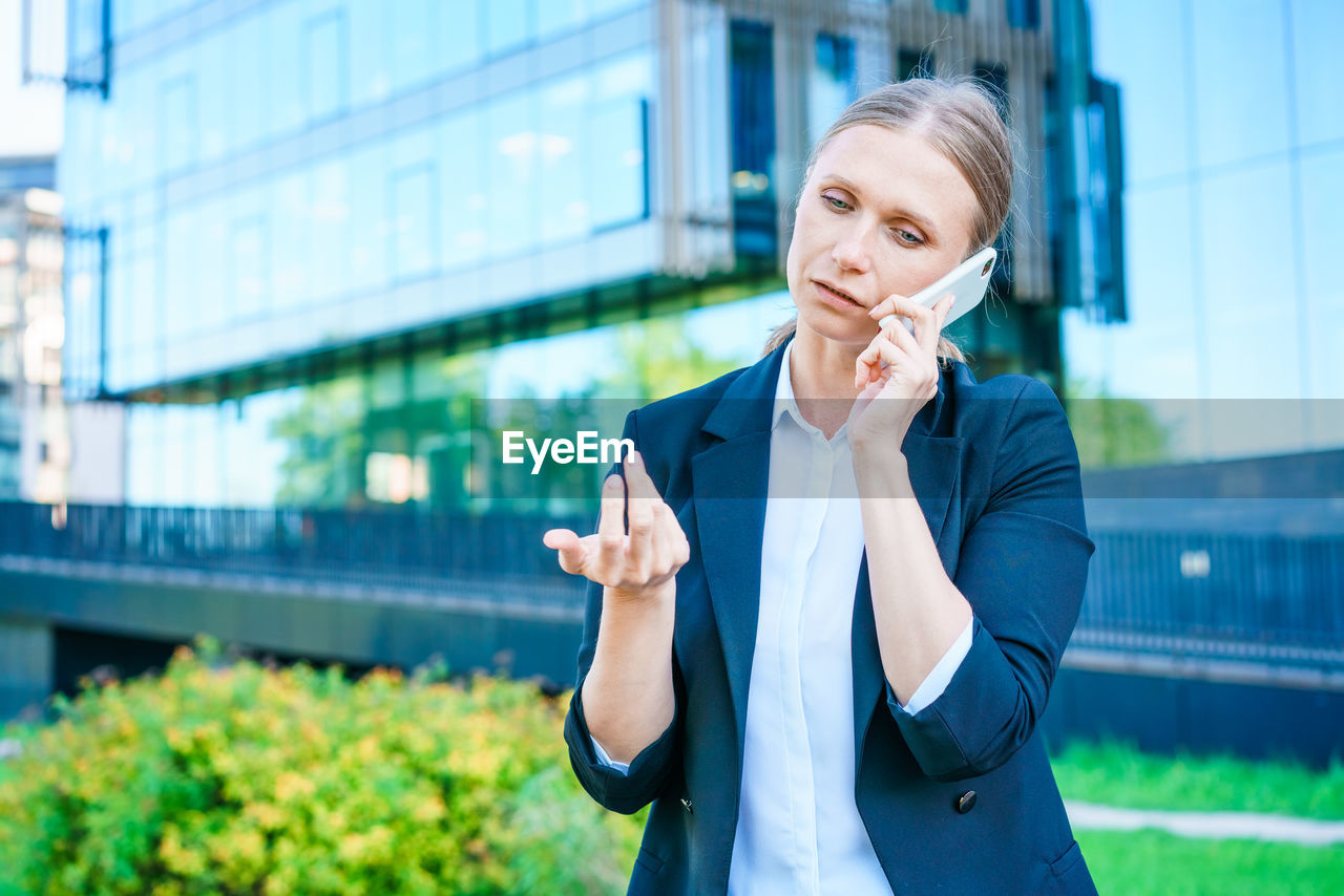 Successful female banker using smart phone outdoors while standing near office