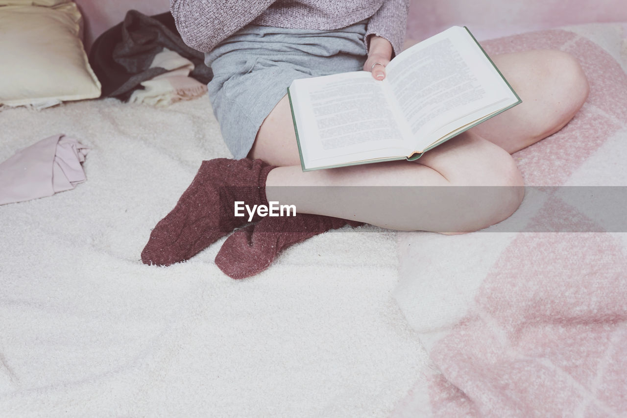 Low section of woman reading book while sitting on bed