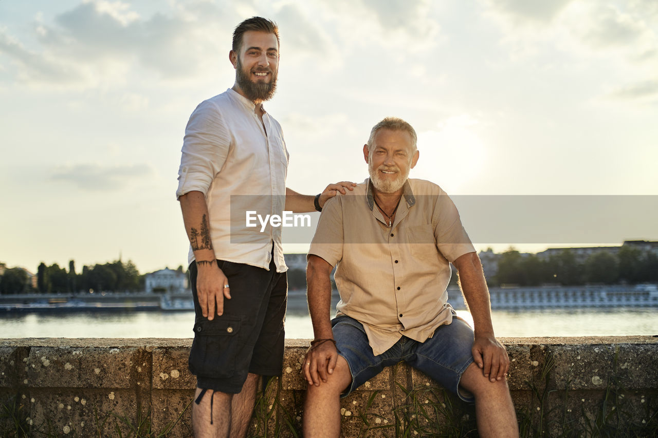 Portrait of smiling father and adult son at the riverside at sunset