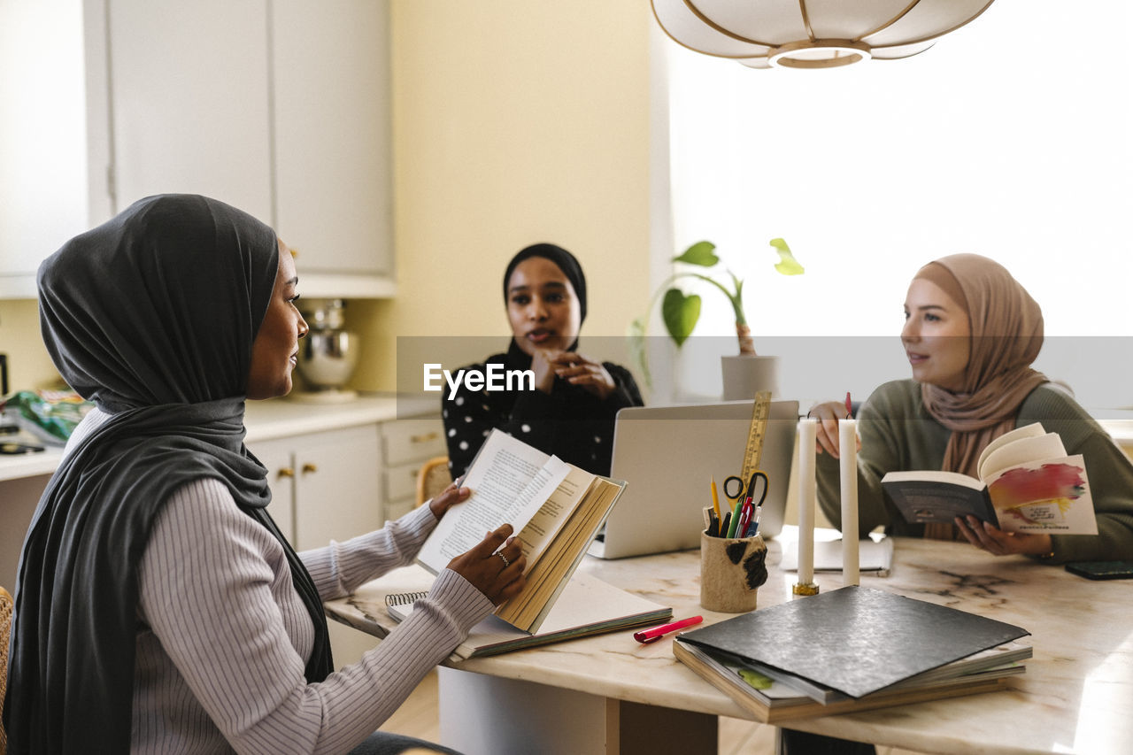 Young woman with female friends reading books while studying together at home
