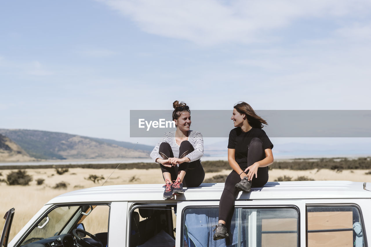 Women sitting on car roof against sky