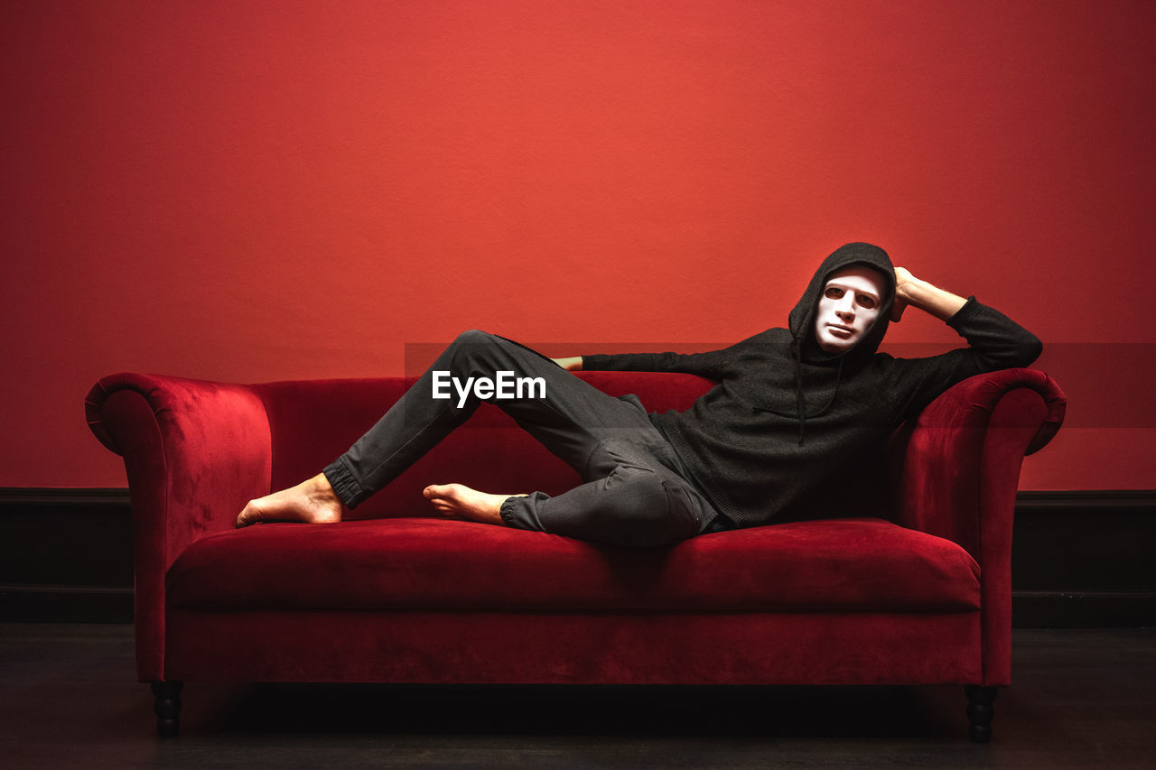 Full length portrait of man wearing spooky mask relaxing on sofa