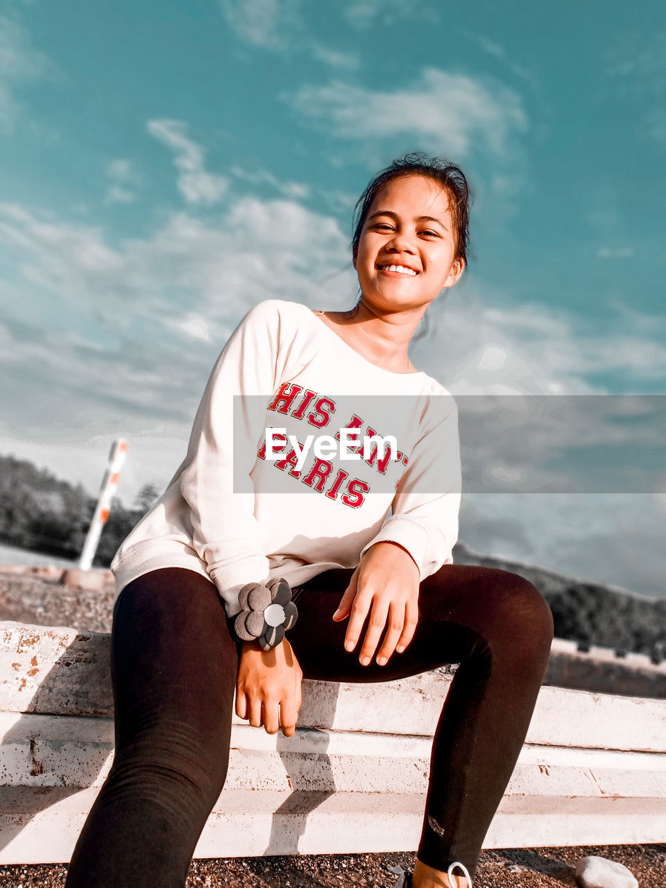 FULL LENGTH OF A SMILING YOUNG WOMAN SITTING AGAINST WATER