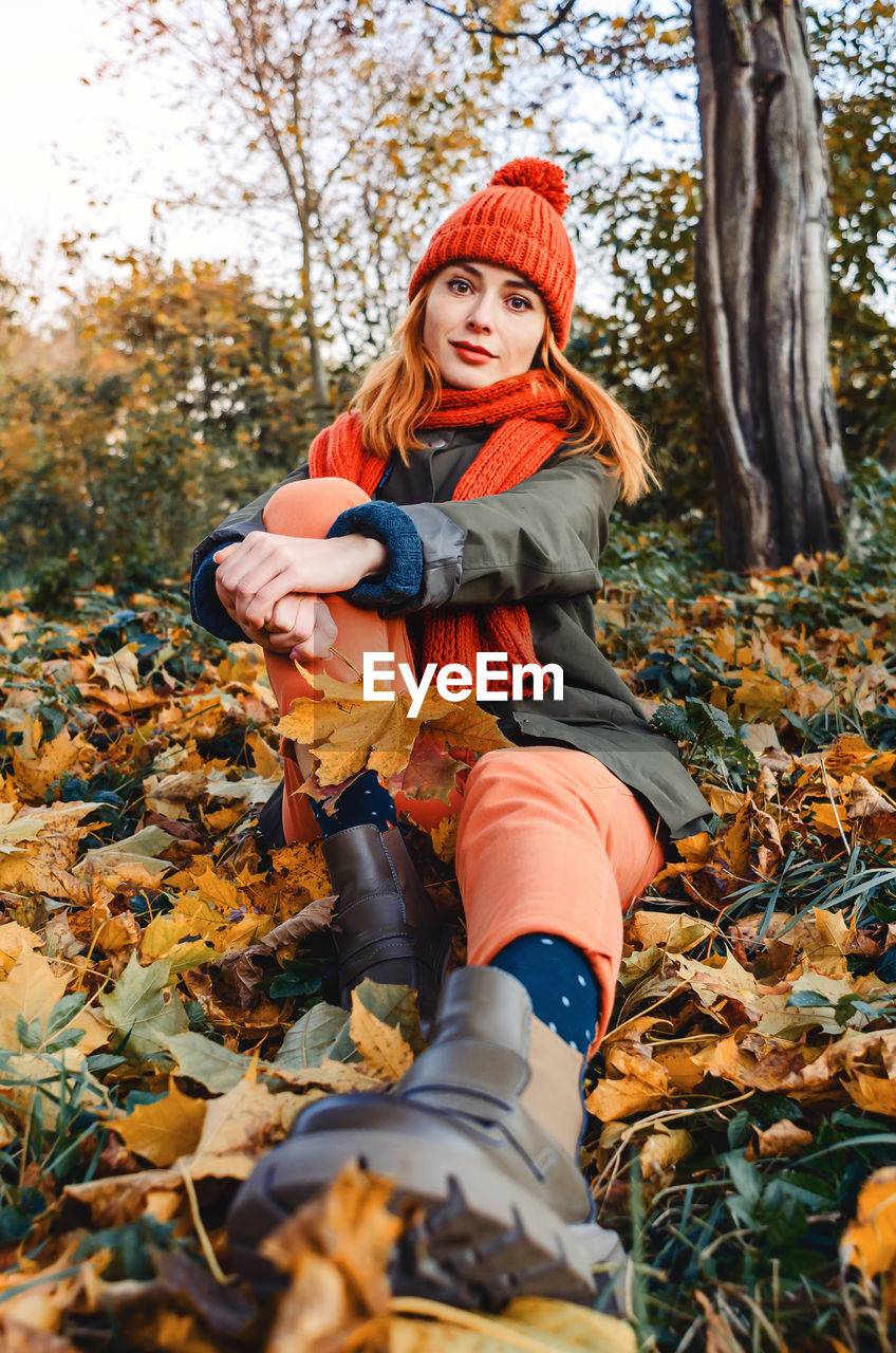 Bright cute young woman in orange warm knitted hat and scarf on background of yellow autumn leaves