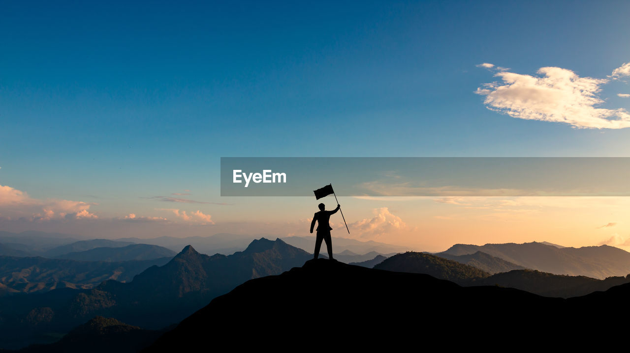 Silhouette man holding flag while standing on mountain peak against sky during sunset