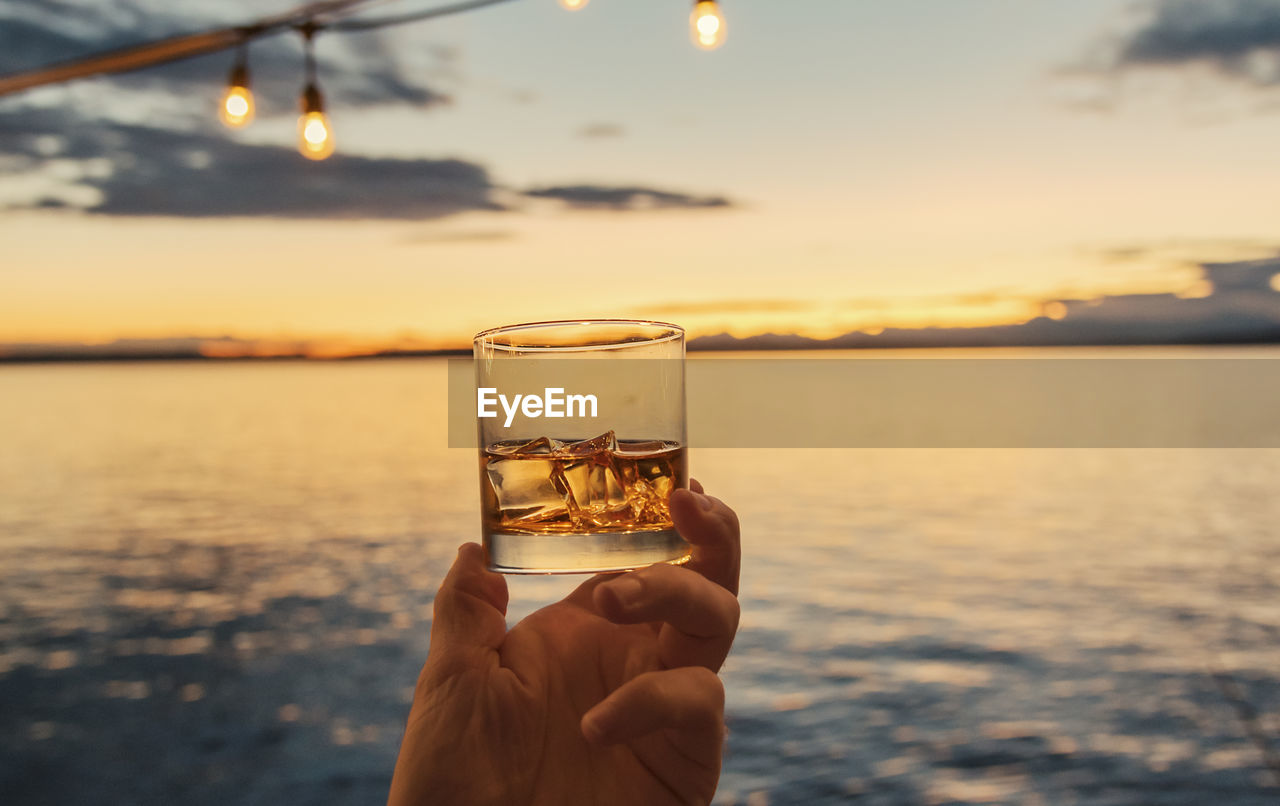 Close-up of hand holding whiskey glass against sea during sunset