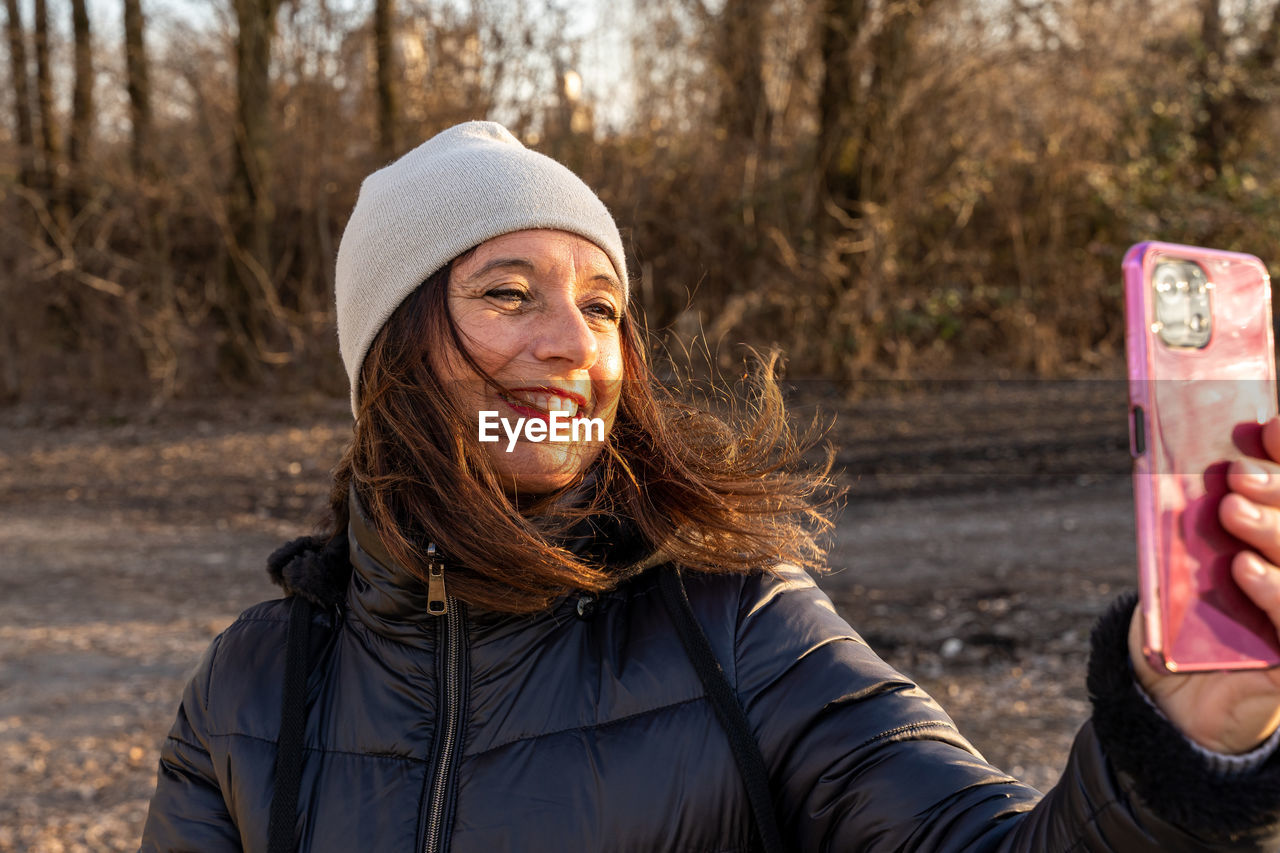 Middle aged woman wearing winter clothes taking selfie by a river - concept of people in recreation