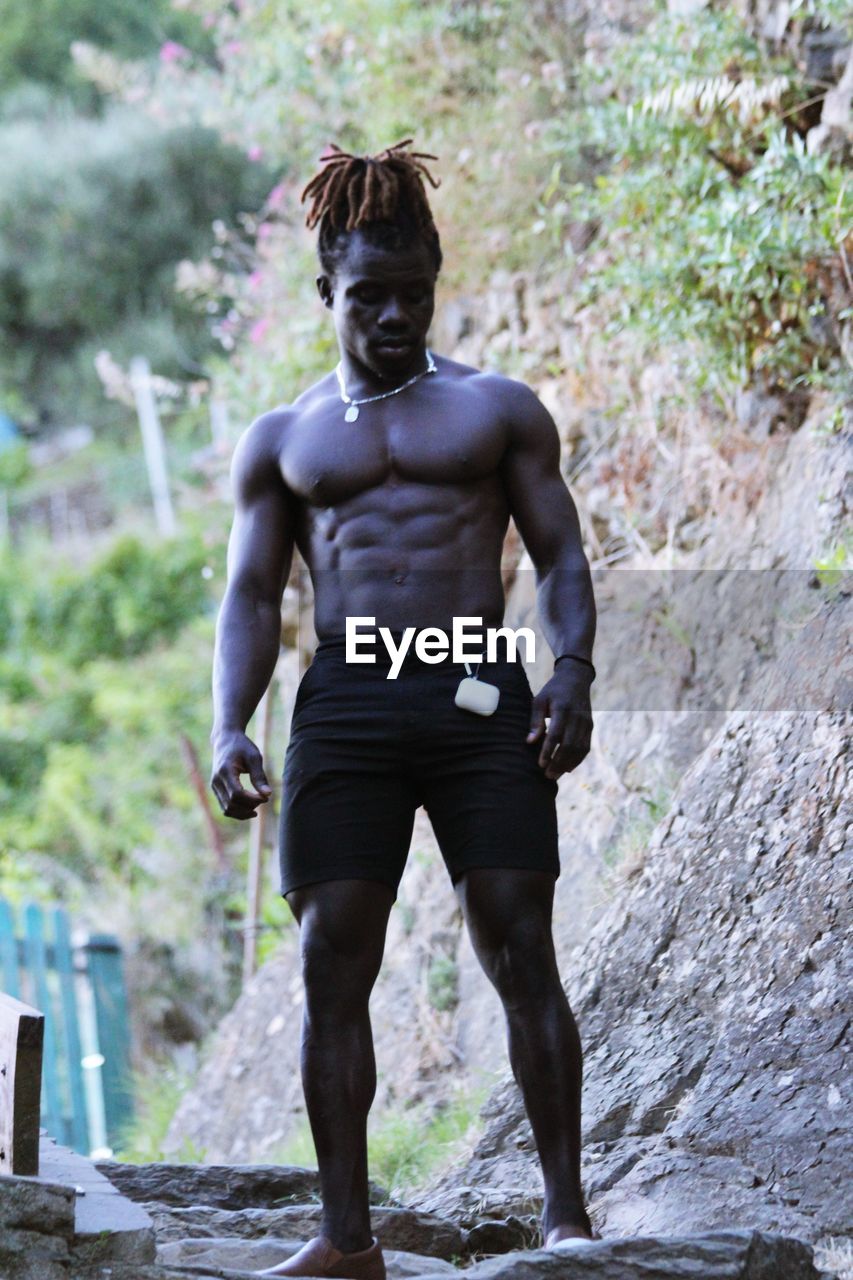 FULL LENGTH OF SHIRTLESS YOUNG MAN STANDING AGAINST WATER