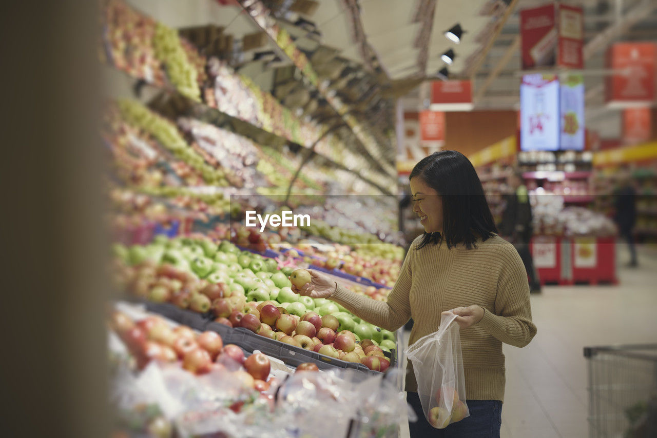 View of woman standing in supermarkets and putting apples into plastic bag