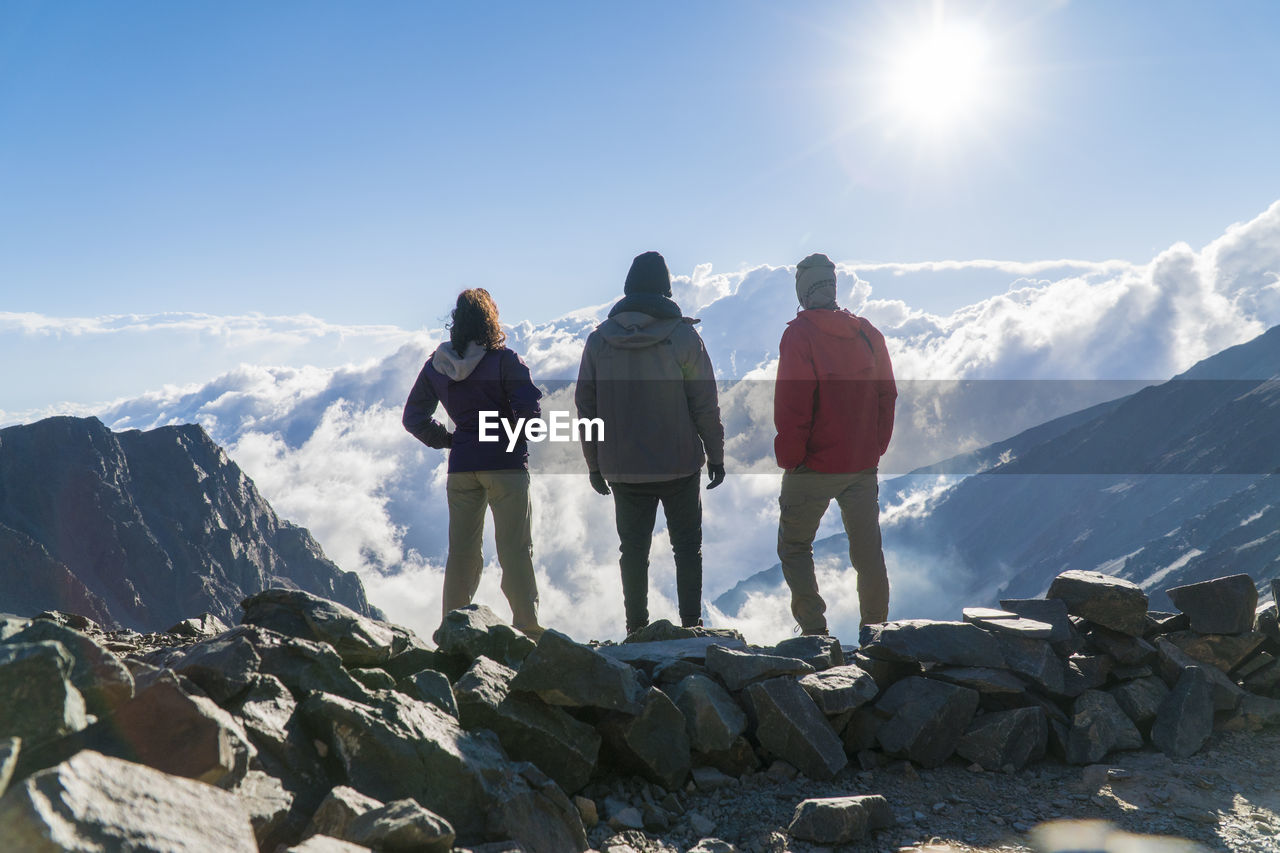 REAR VIEW OF PEOPLE STANDING ON ROCKS AGAINST MOUNTAINS