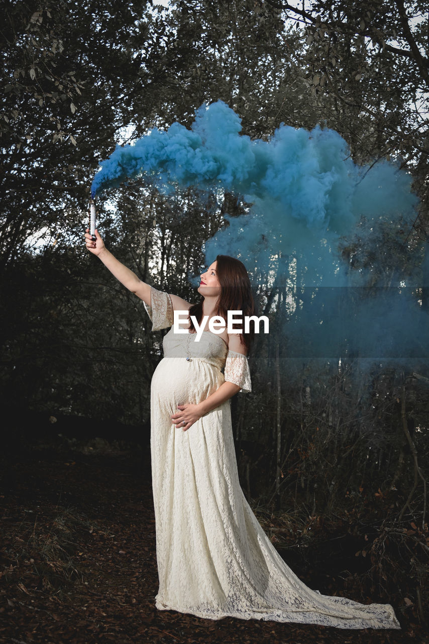 Pregnant woman holding distress flare while standing against trees in forest