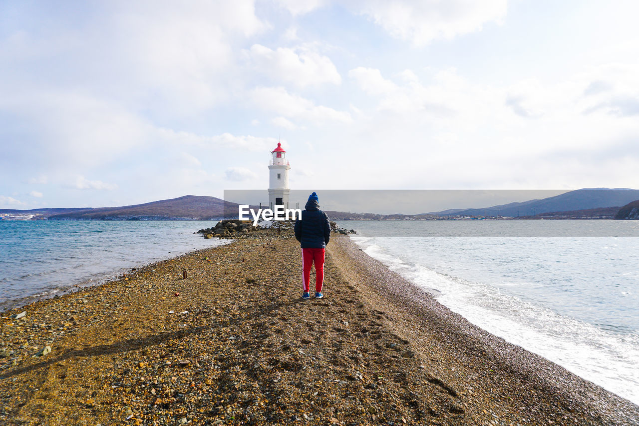 REAR VIEW OF PERSON STANDING ON BEACH AGAINST SKY