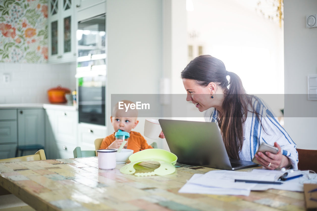 Smiling working mother talking with daughter while freelancing at dining table in kitchen