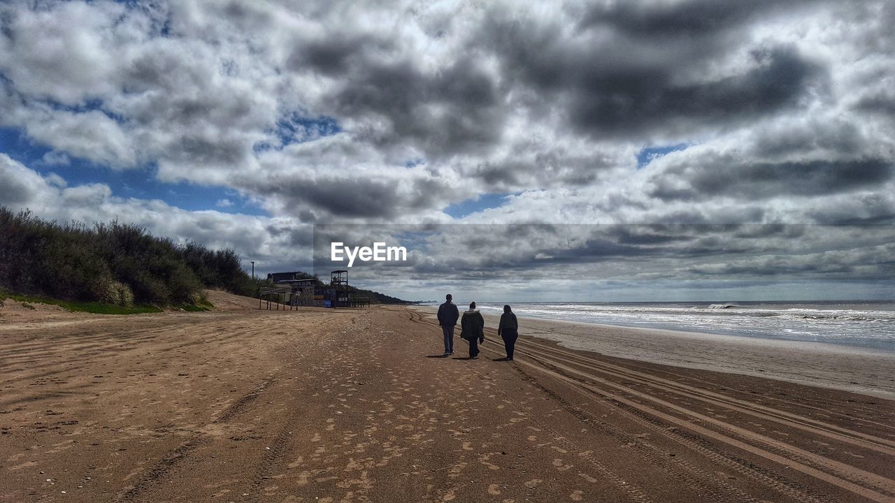 REAR VIEW OF MAN WALKING ON BEACH AGAINST SKY