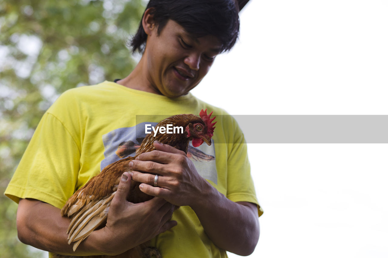 Low angle view of farmer holding hen while standing against clear sky