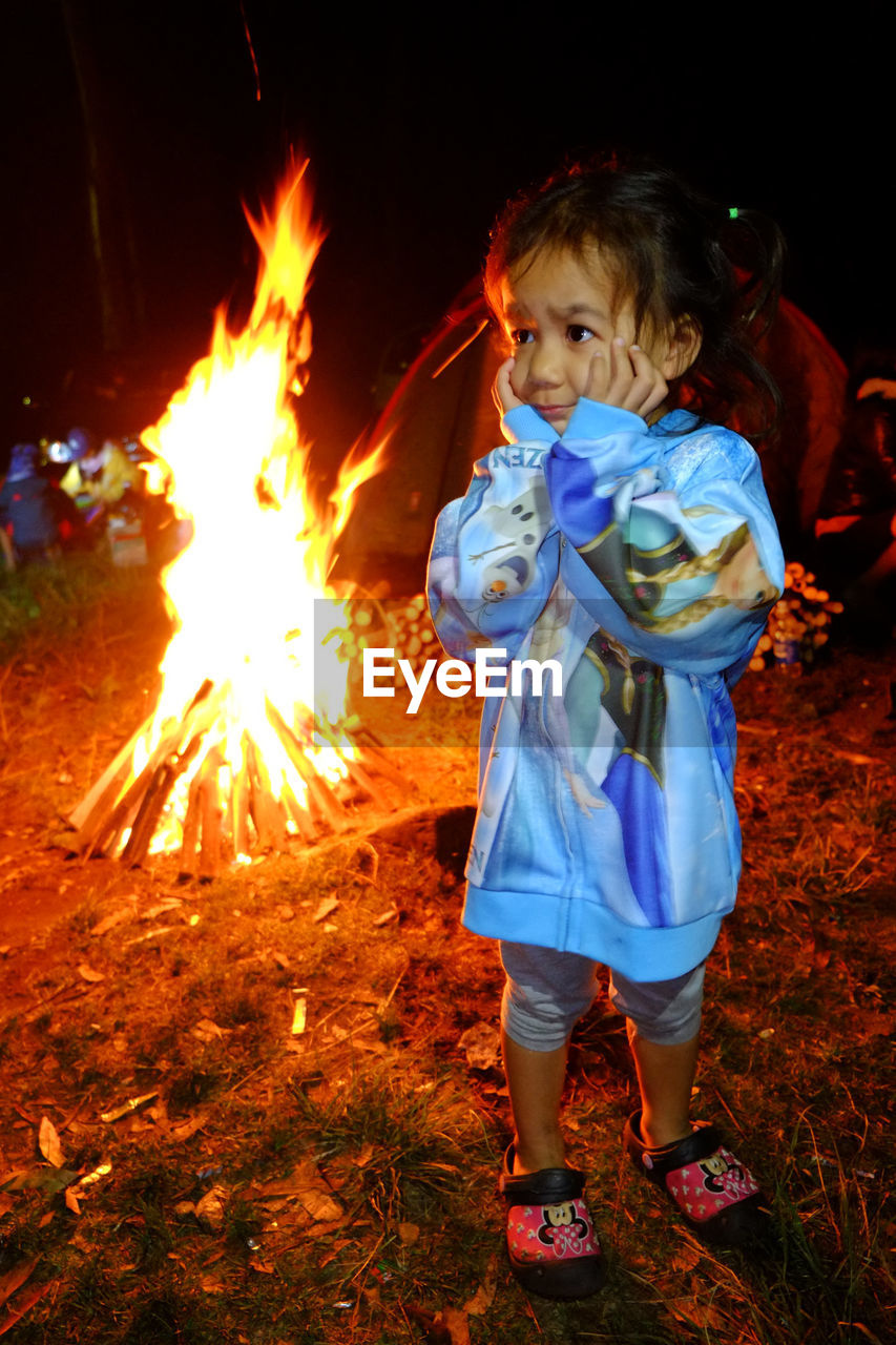 Cute girl with hand on chin standing against campfire on field at night