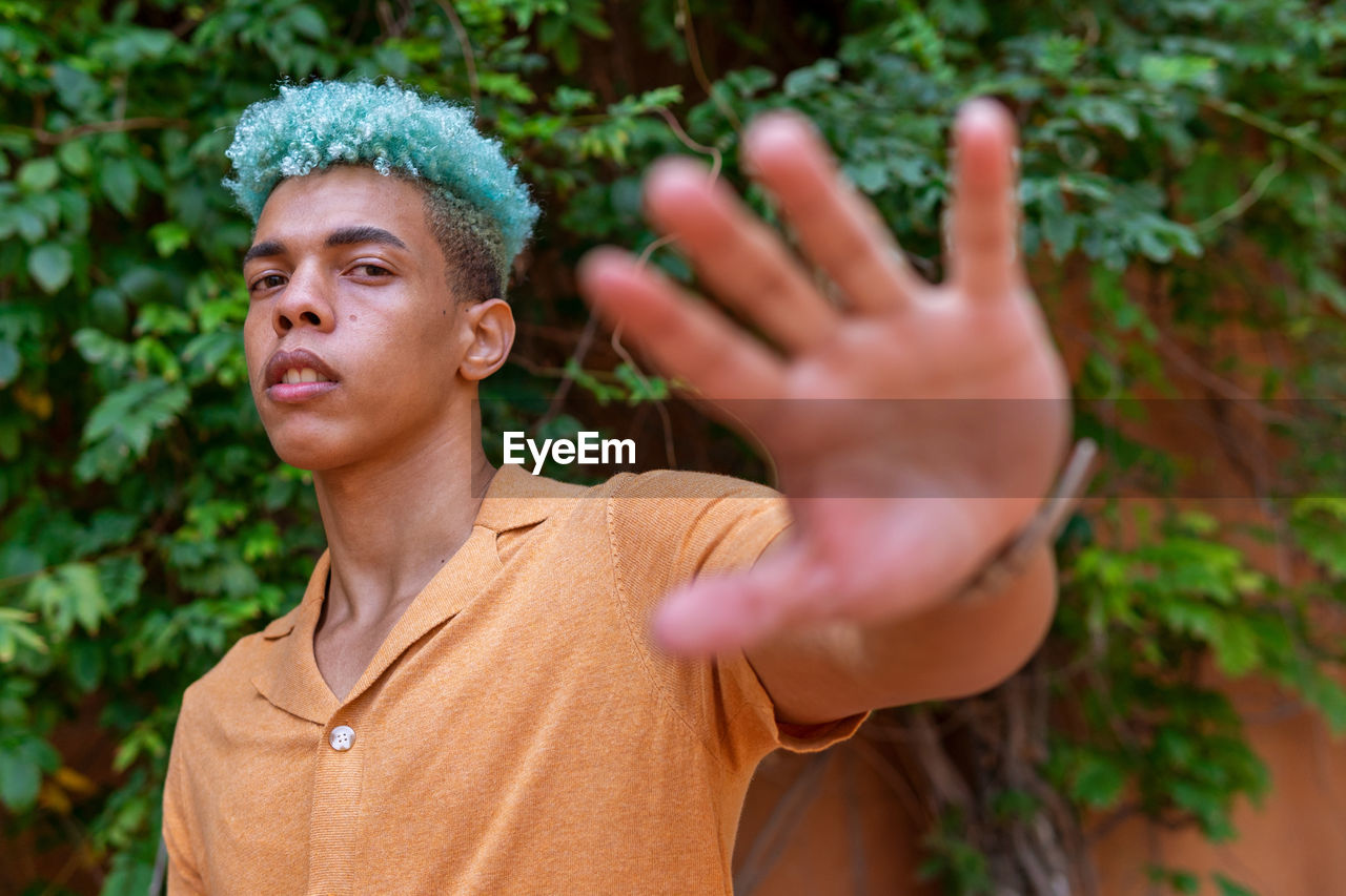 Confident african american young male hipster with blue curly hair reaching hand towards camera while standing on street