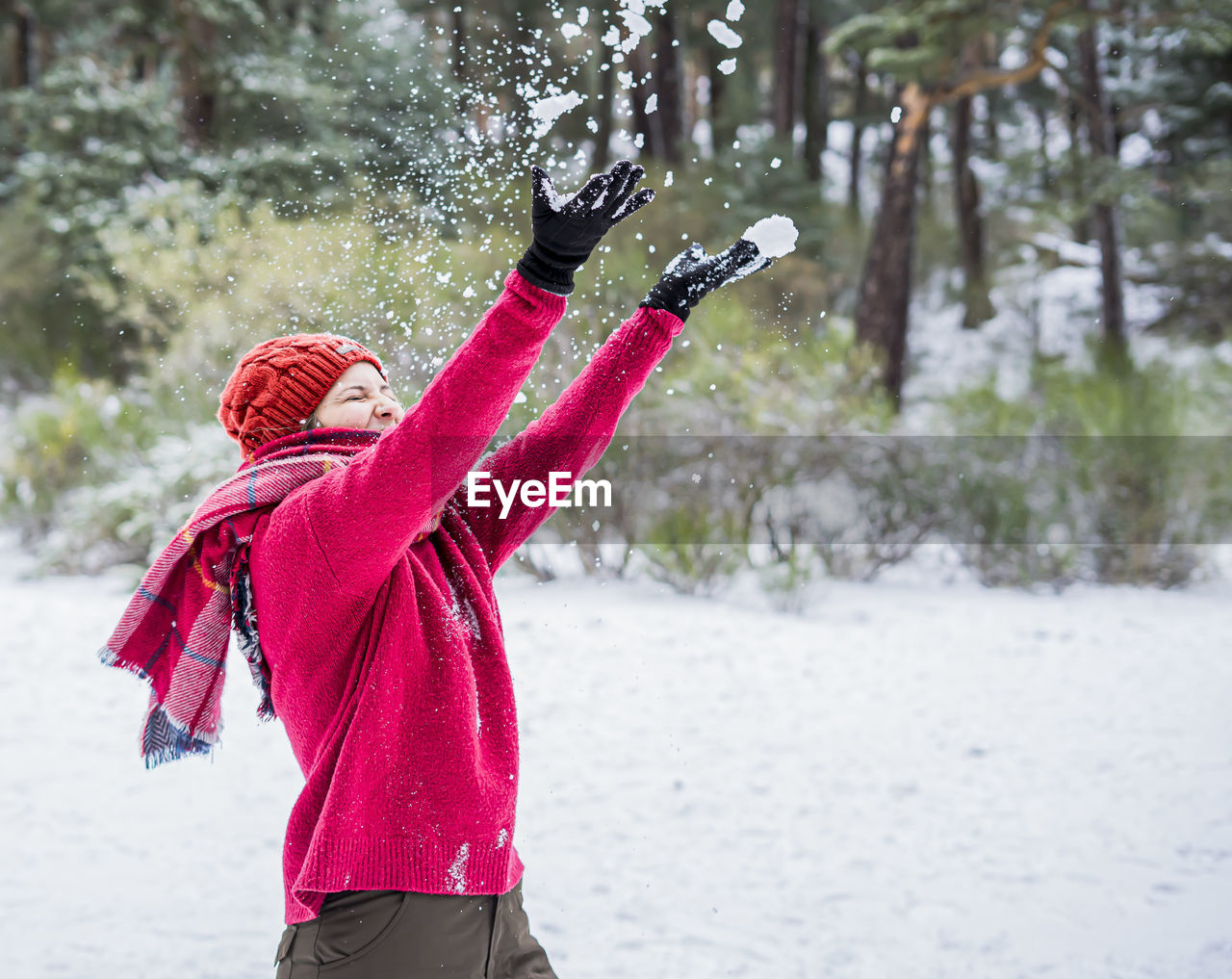 Cheerful young woman throwing snow mid air while standing on snow covered land